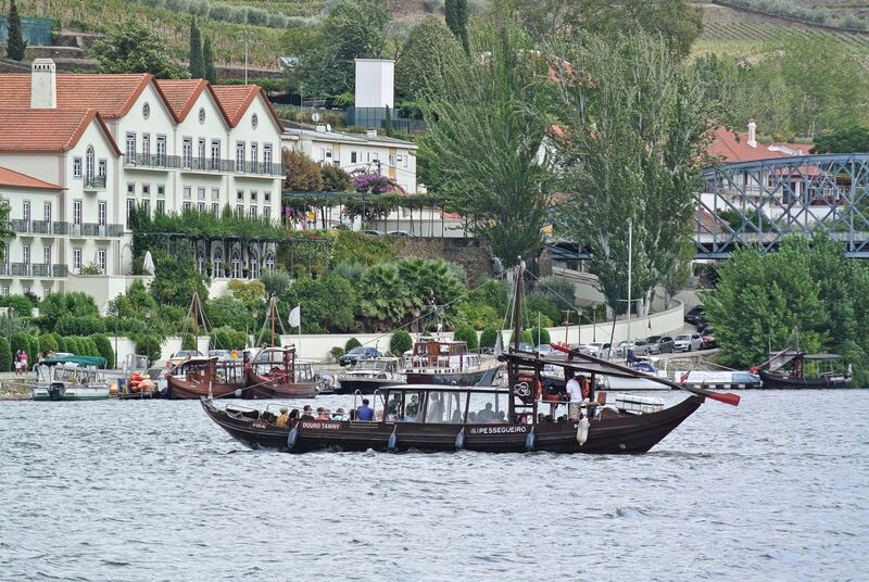 DC 22-0797-070.jpg :: Saga Douro River Cruise, Pocinho to Pinhao, River Douro: Rabelo boat in front of the Ponte do Pinhão, traditionally used for shipping port wine from the vineyards in the Alto Douro region to the wine cellars in Vila Nova de Gaia/Porto. They are characterized by a flat bottom and a long steering oar, designed for navigating the shallow and sometimes challenging waters of the river before modern dams were built. Today, these boats are primarily used for scenic tourist cruises, offering visitors a relaxing way to view the stunning landscapes and historic wine estates along the river, particularly around towns like Pinhão. Photo taken 02/09/2022