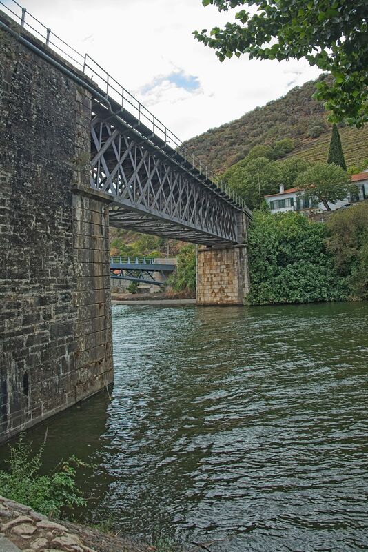 DC 22-0797-109.jpg :: Saga Douro River Cruise, Pocinho to Pinhao, River Douro: The Pinhão Railway Bridge, a prominent truss bridge that spans the Pinhão River at its confluence with the Douro River in Portugal. The structure is part of the historic Linha do Douro (Douro Line), which is renowned for its scenic views of the UNESCO-listed Douro Valley wine region. The bridge features a classic metal lattice truss design supported by robust stone masonry piers that extend into the water. It serves the Pinhão railway station, located nearby, which is famous for its traditional blue-and-white tile (azulejo) panels depicting the region's viticulture. Photo taken 02/09/2022