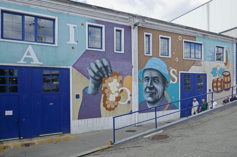 DC 22-0808-063.jpg :: Saga Douro River Cruise, Favaios:  The building in the image is the Adega Cooperativa de Favaios, a renowned winery located in the village of Favaios within the Douro wine region of Portugal. The winery is famous for producing one of Portugal's most iconic fortified wines, Moscatel de Favaios. The winery is nestled in the picturesque Alto Douro wine region of Portugal, a UNESCO World Heritage site. Founded in 1952, the cooperative was established by local producers to create their famous Moscatel wine. While most famous for its Moscatel, the winery also produces DOC Douro wines (white, red, and rosé), sparkling wines, and Port wine. This vibrant street art is a mural on the building of the Adega Cooperativa de Favaios. The mural features a portrait of a local figure known as \