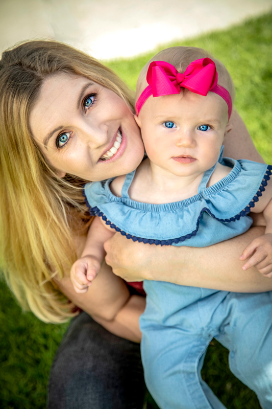 Close up of blonde woman and her baby girl 001A.jpg :: Cute close up of blonde mother with blue eyes holding her daughter with blue eyes and pink bow and both looking up at the camera while sitting at a park on green grass