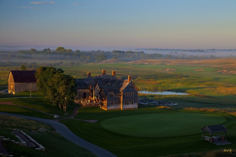 E101--0161  V2  1500px  SS  with signature1.jpg :: The Lodge, practice putting green, starter's canopy, original barn and outdoor terrace with the par five first hole coming in from the center right and extending around the wetland and above the Lodge.  In the lower right corner is the stairway leading from the Lodge up to the Village Clubhouse and the cabins.  Erin Hills is a daily fee golf course in Erin, Wisconsin and was designed by Hurdzan/Fry and Ron Whitten and built by Landscapes Unlimited, LLC, and has been choosen to host the 2011 US Amateur Chanpionship and the 2017 US Open Championship.  Photo by Paul Hundley; taken in August, 2012.