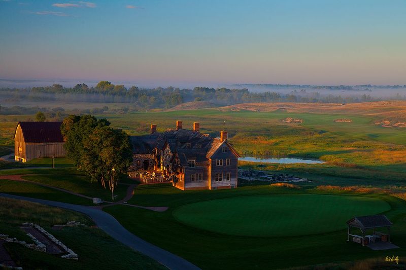 E101--0161  V2  1500px  SS  with signature.jpg :: The Lodge, practice putting green, starter's canopy, original barn and outdoor terrace with the par five first hole coming in from the center right and extending around the wetland and above the Lodge.  In the lower right corner is the stairway leading from the Lodge up to the Village Clubhouse and the cabins.  Erin Hills is a daily fee golf course in Erin, Wisconsin and was designed by Hurdzan/Fry and Ron Whitten and built by Landscapes Unlimited, LLC, and has been choosen to host the 2011 US Amateur Chanpionship and the 2017 US Open Championship.  Photo by Paul Hundley; taken in August, 2012.